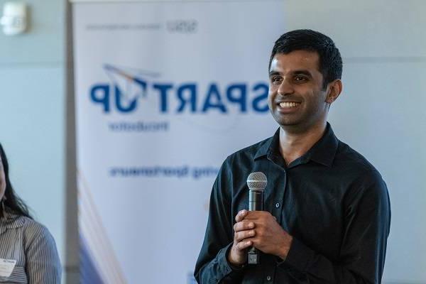 A dark-haired man holds a microphone and smiles, with a white pop-up banner that says "SpartUp Incubator" behind him.